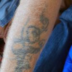 Foto av översida underarm med gråblek tatuering föreställande gissningsvis snirklig blomma med kringliggande, spiralformadbanderoll.