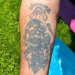 Foto av underarm med äldre, något grumlig tatuering. Tatueringen föreställer två skepp i överliggande formation, delade i två bildramar av vågor och banderoll som löper över tatueringens mitt, mellan de båda skeppen. Text och molnkonturer ramar in det hela. Längst ned syns ett ankare.