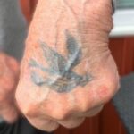 Foto av handrygg med urblekt, svartfrå tatuering föreställande en flygande svala med ett kuvert och en banderoll i näbben. I banderollen står ett numera oläsligt namn.