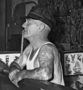 Svartvitt porträttfoto av August Cap Coleman. Rakat huvud, klädd i vegamössa. Tatuerade armar.