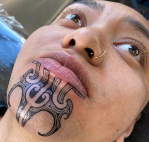 Färgfotografi av en maorisk kvinnas ansikte i närbild. Hennes haka pryds av en traditionell maorisk tatuering i svart.