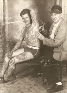 Svartvitt fotografi av två män där den ene tatuerar den andre på ryggen. Mannen som tatueras är den tatuerade mannen Tom Stephens. Tatueraren är Bert Grim. Båda herrarna har signerat fotot.