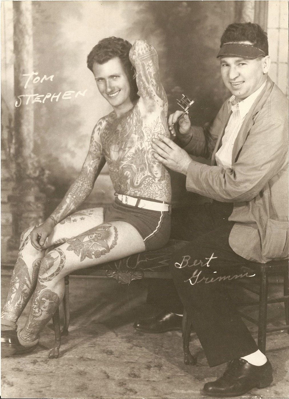 Svartvitt fotografi av två män där den ene tatuerar den andre på ryggen. Mannen som tatueras är den tatuerade mannen Tom Stephens. Tatueraren är Bert Grim. Båda herrarna har signerat fotot.