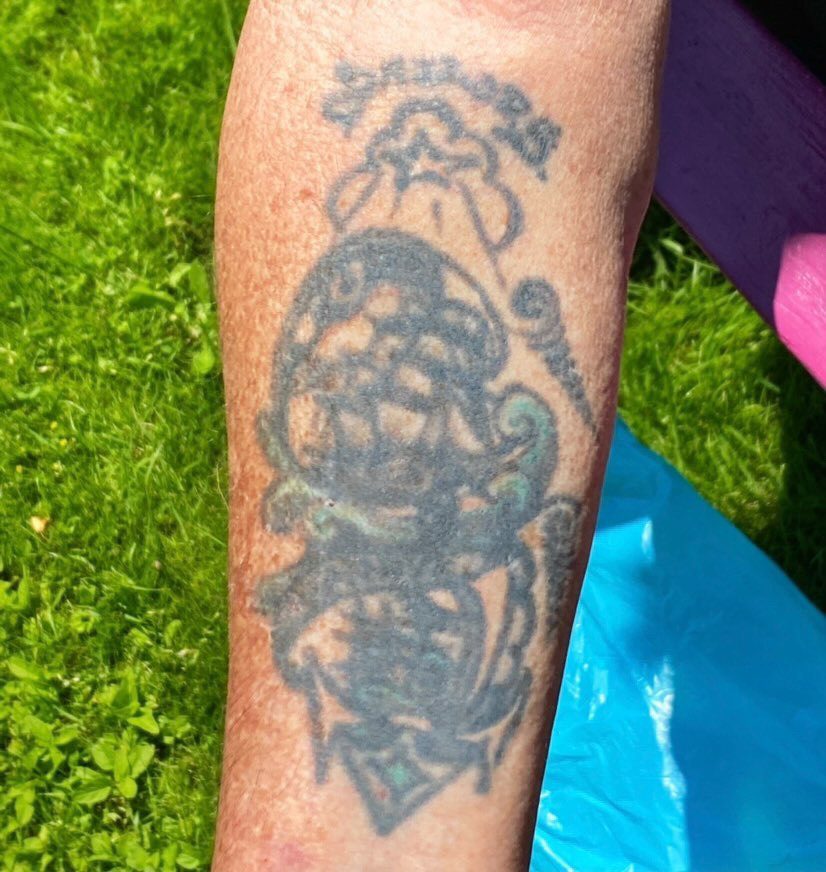 Foto av underarm med äldre, något grumlig tatuering. Tatueringen föreställer två skepp i överliggande formation, delade i två bildramar av vågor och banderoll som löper över tatueringens mitt, mellan de båda skeppen. Text och molnkonturer ramar in det hela. Längst ned syns ett ankare.