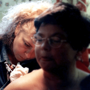 Färgfoto av Mia Nén, sittandes framåtlutad mot en kvinnan rygg medan hon tatuerar kvinnan på skuldran.