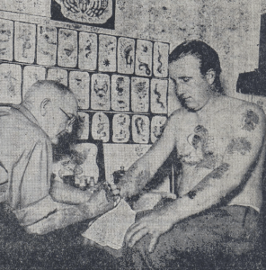 Raserat, svartvitt foto från äldre tidningsartikel. En man i glasögon och skjorta med uppkävlade ärmar sitter framåtböjd och tatuerar en yngling med tatuerad, bar överkropp på höger underarm.