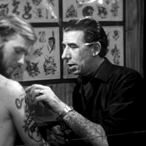 Svartvit foto av Tato Jim från danskt TV-inslag, 1965. Mager man med bakåtkammat hår och långa, grova ansiktsdrag och skarpa ögonbryn sitter och petar på en tatuering på en man, blond man i bar överkropp.