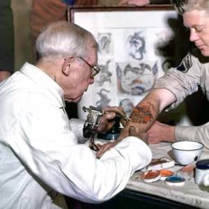 Färgfoto av George Burchett – en liten äldre tunnhårig grå man, iklädd vit läkarrock och svarta runda glasögon, tatuerandes en ung flottist på under armen.