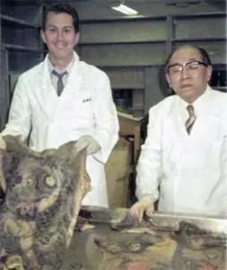 Iklädda vita läkarrockar, framför ett stålbord med utskurna och konserverade, tatuerade människohudar, står en ung Ed Hardy tillsammans med Masaichis son, doktor Fukushi Katsunari. Hardy håller leende upp exemplaret av en tatuerad ryggtavla.