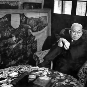 Svartvitt foto av doktor Fukushi Masaichi. Äldre japansk man med vitt hår och mustasch iklädd svart kostym, sitter avslappnat i fåtölj. I bakgrunden syns en stor ram där en stor bit rygghud med en japansk ryggtatuering finns inglasad. På bordet framför mannen ligger mindre tatuerade hudbitar och foton utbredda.