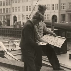 Svartvitt foto av två män intill en kanal där en båt ligger förtöjd. Ena mannen håller upp ett ark med tatueringsmotiv. Tatuerare Holger Jørgensen går omkring i Nyhavn för att göra reklam för sin firma.