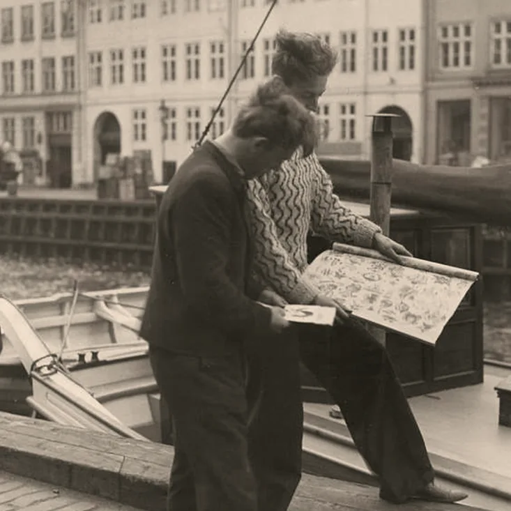 Svartvitt foto av två män intill en kanal där en båt ligger förtöjd. Ena mannen håller upp ett ark med tatueringsmotiv. Tatuerare Holger Jørgensen går omkring i Nyhavn för att göra reklam för sin firma.