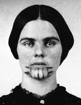 Svartvitt närbildsporträtt av Olive Oatman. Ett i efterhand färglagt porträttfoto av Olive Oatman. På bilden syns en ljushyllt ung kvinna med mörktbrunt hår i tidenlig (amerikanskt 1860-tal) håruppsättning. På hennes haka syns en tatuering i form av fem lodräta linjer från underläppen till haklinjen. De yttersta linjerna som går ned från mungiporna pryds av fyra – två på vardera sidan, koniska taggar som pekar ut mot kinderna.