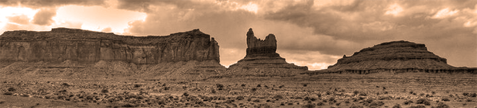 Ökenlandskap, Arizona, USA. Skymningsbild av torrt, karg ökenlandskap med låga tistel- och kaktusväxter. Vid horisonten syns höga, rektangulära och kvadratformade klippmassiv. Himlen bortom bergen består av ljusstrimmade moln.