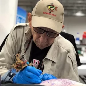 Färgfoto av Bowery Stan i arbete. En äldre man i beiga kläder sitter framåtlutad över att axelparti som syns i förgrunden, på vilket han tatuerar. Stans händer är klädda i blå skyddshandskar, tatueringsmaskinen är brons- och guldfärgad.