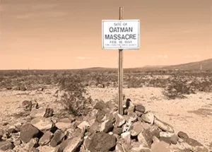 Foto av minnesplatsen för familjen Oatman. I ett ökenlandskap står en plåtskylt i ett stenröse. På skylten står att läsa: Site of Oatman Massacre. Feb 18 1851. Yuma County Historical Society.