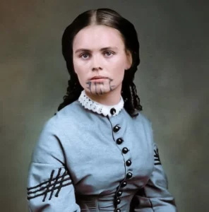 Ett i efterhand färglagt porträttfoto av Olive Oatman. På bilden syns en ljushyllt ung kvinna med axellångt mörktbrunt hår i tidenlig (amerikanskt 1860-tal) håruppsättning. Hon är klädd i en ljusblå klänning med vit krage och svarta runda knappar över bröstet. På hennes haka syns en blågrön tatuering i form av fem lodräta linjer från underläppen till haklinjen. De yttersta linjerna som går ned från mungiporna pryds av fyra – två på vardera sidan, koniska taggar som pekar ut mot kinderna.