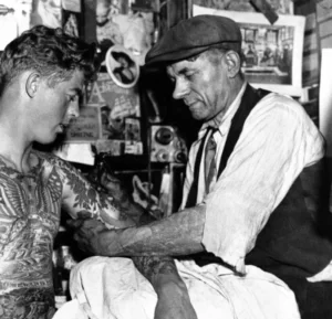 Svartvitt foto av Charlie Wagner tatuerandes ung kund. Till vänster i bild sitter en ung man i bar överkropp. Bröst, mage och armar är tatuerade. Till höger sitter en man i vit skjorta, vitt förkläde, märk väst och 1930-talskeps hållandes i unge kundens vänstra arm med sin vänstra hand, medan höger hand håller i en tatueringsmaskin med vilken kundens vänstra överarm tatueras.
