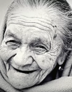 Svartvitt porträttfoto av äldre kvinna som med brett leende och varm blick betraktar kameran.