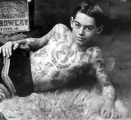 Svartvitt foto av ung Andrew Andy Stuertz. En mörkhårig pojke med bar, tatuerad överkropp ligger på sidan på en djurfäll, stödjandes på ena armbågen. Han tittar spjuveraktigt rakt in i kameran. Den andra armen stöds slappt med handen mot ena höften.